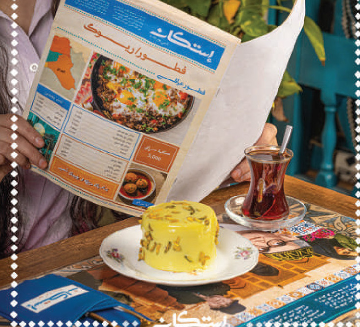 Guest reading the Estikan newspaper-style menu with tea and a saffron cake