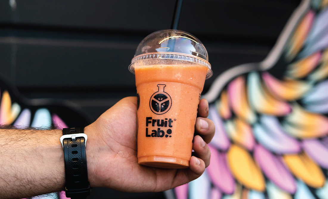 Hand holding an orange Fruit Lab smoothie cup in front of a wing mural